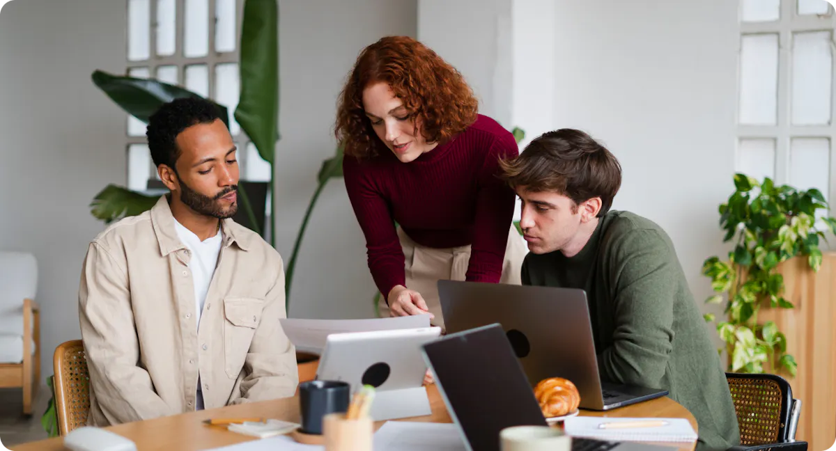 Three colleagues reviewing printed documents together at a table with laptops
